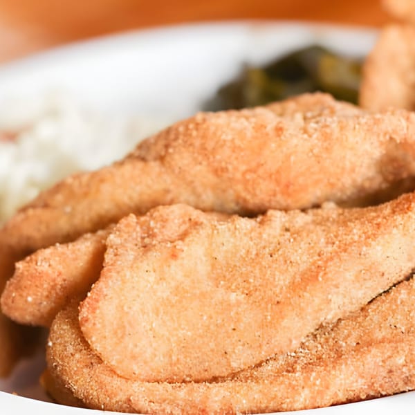 Close-up of golden-brown fried catfish fillets with sides of coleslaw and collard greens.