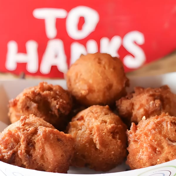 Golden-brown hush puppies in a white paper container, with a red Hank's sign blurred in the background.